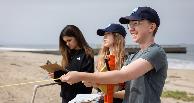 Three LMU students measuring the beach and holding clipboards